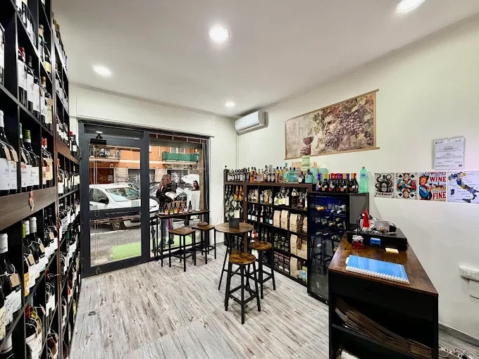 Interno dell'Enoteca Per Bacco: scaffali scuri pieni di bottiglie, tavolini alti e sgabelli, quadro con uva