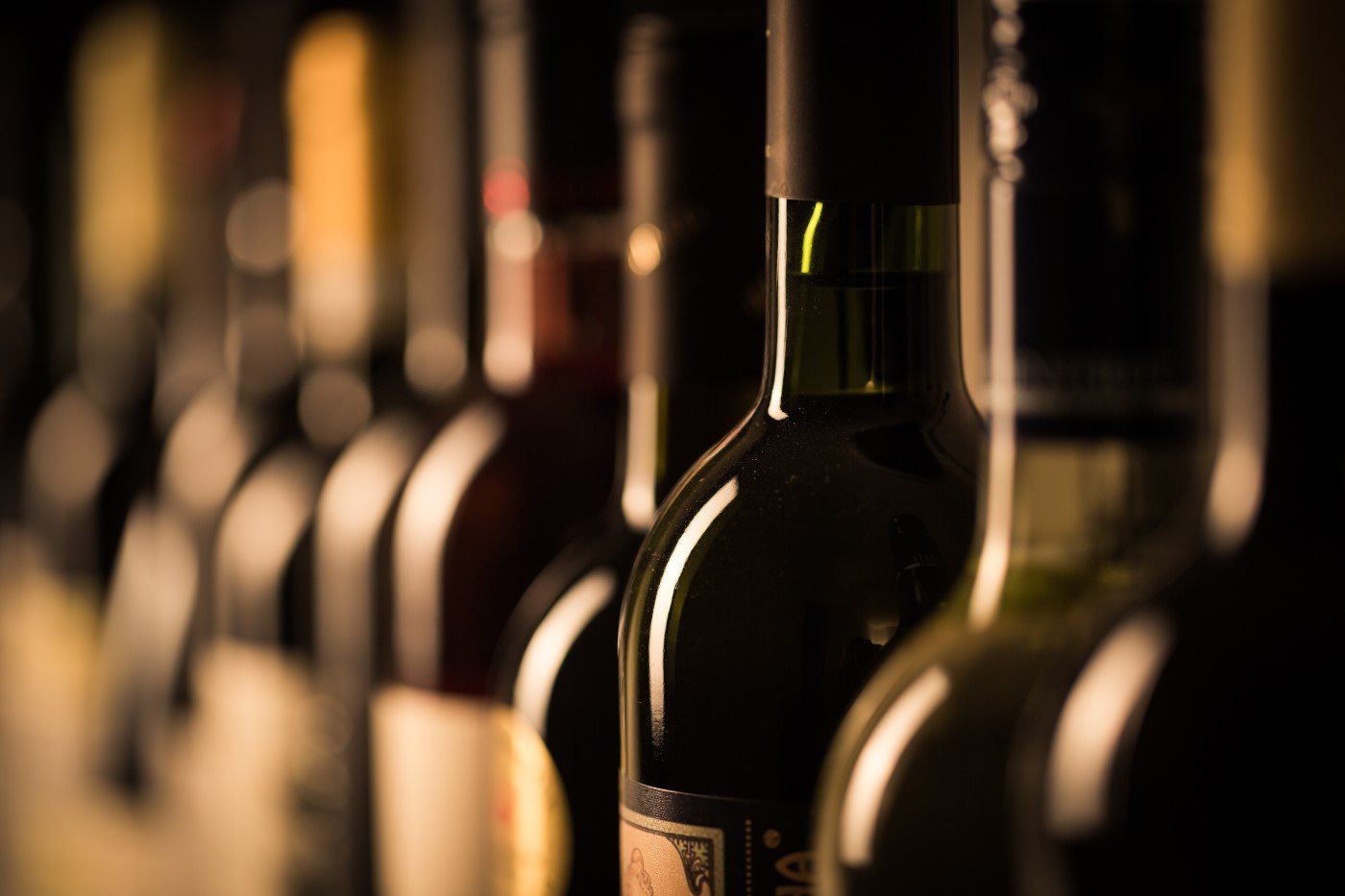 Fila di bottiglie di vino scuro con capsule nere, in primo piano un collo di vetro verde illuminato da luce calda