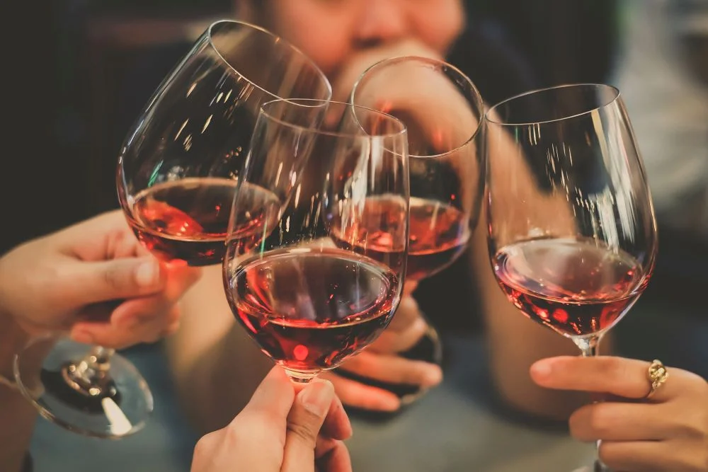 Quattro mani che brindano con calici di vino rosso su sfondo sfocato, una con un anello dorato al dito
