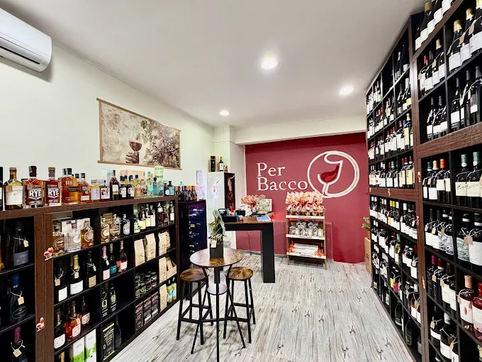 Interno con scaffali di legno pieni di vini e liquori, tavolino alto con sgabelli e parete rossa con logo Per Bacco
